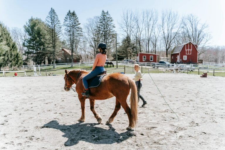A woman learning horseback riding in an outdoor arena with instructor guidance.