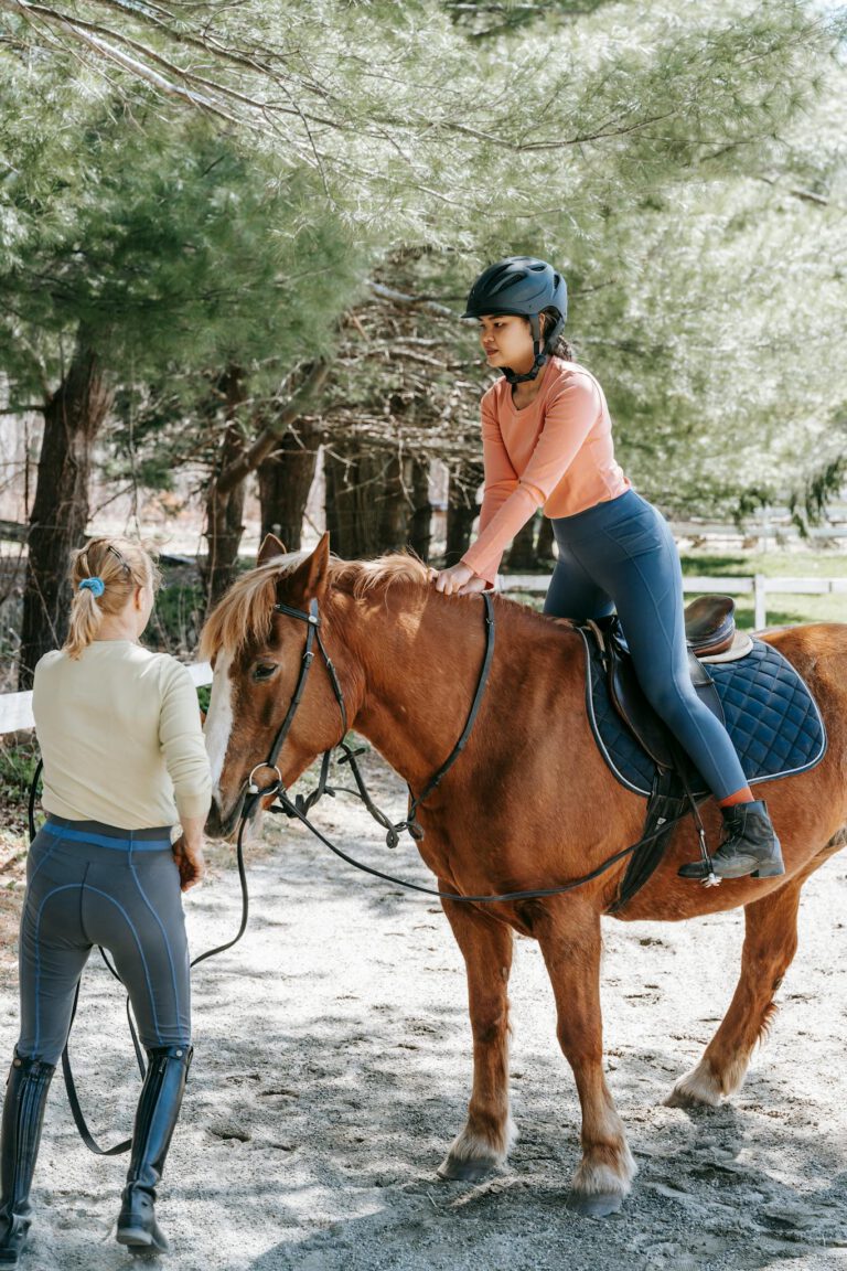 Woman learning horseback riding with instructor in a forest setting. Clear day outdoors.